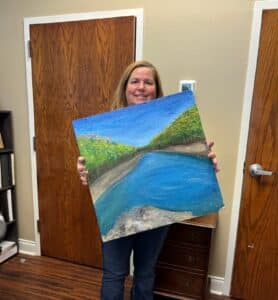 Woman holding a painted ceiling tile with a lakeside image